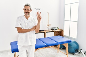 Fototapeta na wymiar Middle age hispanic therapist man working at pain recovery clinic smiling with happy face winking at the camera doing victory sign with fingers. number two.