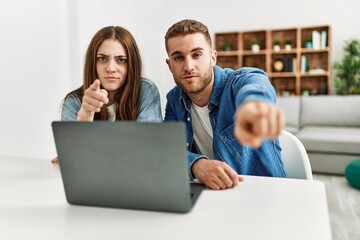 Young caucasian couple working using computer laptop at home pointing with finger to the camera and to you, confident gesture looking serious