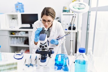 Young hispanic woman wearing scientist uniform using microscope at laboratory