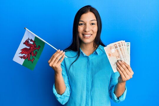 Young latin woman holding wales flag and pounds banknotes smiling with a happy and cool smile on face. showing teeth.