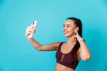 Music For Workout. Portrait Of Smiling Young Slim Lady Wearing Wireless Earphones, Enjoying Favorite Song And Playlist For Training, Holding Smartphone Isolated Over blue Studio Background