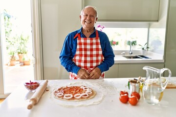 Senior man with grey hair cooking pizza at home kitchen looking positive and happy standing and smiling with a confident smile showing teeth