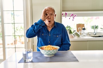 Senior man with grey hair eating pasta spaghetti at home doing ok gesture shocked with surprised face, eye looking through fingers. unbelieving expression.