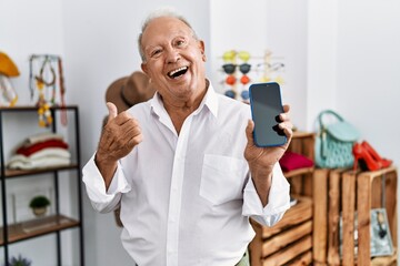 Senior man holding smartphone at retail shop pointing thumb up to the side smiling happy with open mouth