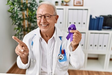 Senior doctor man holding purple ribbon awareness smiling happy pointing with hand and finger to...