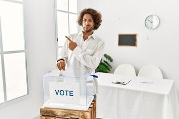 Young hispanic man voting putting envelop in ballot box pointing aside worried and nervous with...