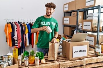 Young arab man wearing volunteer uniform insert food on paper bag at charity center