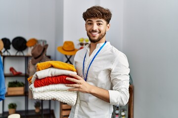 Young arab man shopkeeper holding folded clothes working at clothing store
