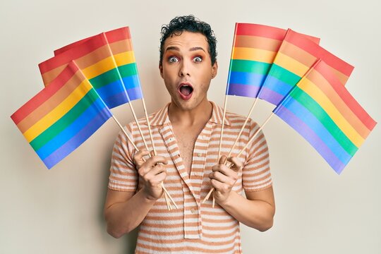 Handsome man wearing make up holding rainbow lgbtq flags afraid and shocked with surprise and amazed expression, fear and excited face.
