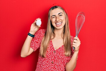 Beautiful hispanic woman holding egg and baker whisk winking looking at the camera with sexy expression, cheerful and happy face.