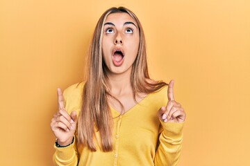 Beautiful hispanic woman wearing casual yellow sweater amazed and surprised looking up and pointing with fingers and raised arms.