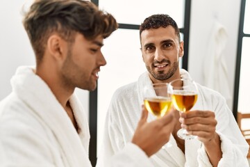 Two hispanic men couple toasting with champagne sitting on massage table at beauty center