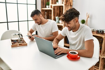 Obraz premium Two hispanic men couple having breakfast using smartphone and laptop at home