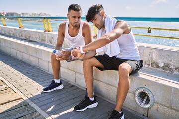 Two hispanic men sporty couple smiling confident looking watch sitting on bench at seaside