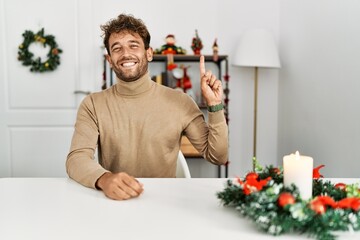 Young handsome man with beard sitting on the table by christmas decoration showing and pointing up with finger number one while smiling confident and happy.