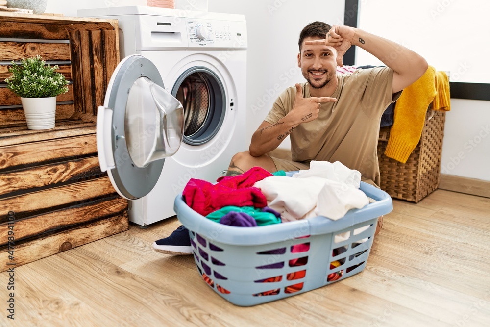 Poster Young handsome man putting dirty laundry into washing machine smiling making frame with hands and fingers with happy face. creativity and photography concept.