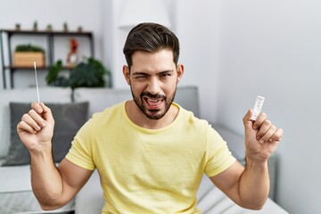 Young man with beard holding coronavirus infection nasal test winking looking at the camera with...