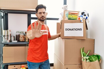 Young man with beard wearing volunteer t shirt at donations stand with open hand doing stop sign...