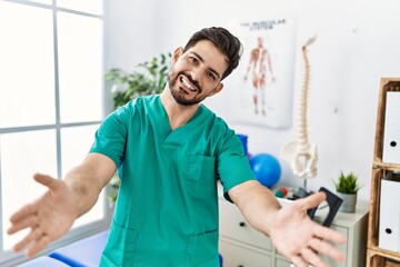 Young man with beard working at pain recovery clinic looking at the camera smiling with open arms for hug. cheerful expression embracing happiness.