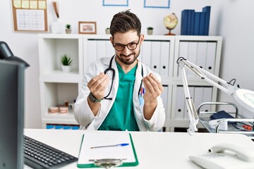 Young man with beard wearing doctor uniform and stethoscope at the clinic doing money gesture with...