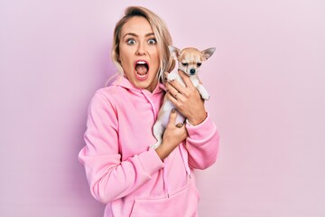 Beautiful young blonde woman hugging cute chihuahua dog afraid and shocked with surprise and amazed...