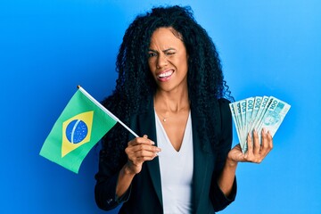Middle age african american woman holding brazil flag and real banknotes winking looking at the...