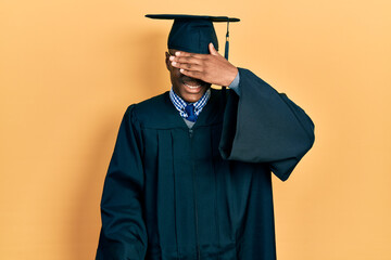 Young african american man wearing graduation cap and ceremony robe smiling and laughing with hand on face covering eyes for surprise. blind concept.