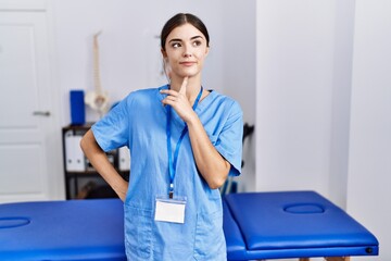 Young hispanic woman wearing physiotherapist uniform standing at clinic thinking concentrated about doubt with finger on chin and looking up wondering