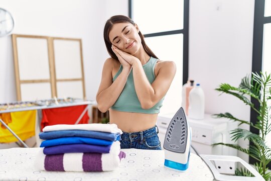 Young hispanic woman ironing clothes at laundry room sleeping tired dreaming and posing with hands together while smiling with closed eyes.