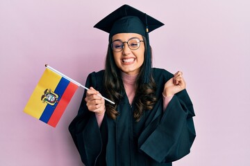 Young hispanic woman wearing graduation uniform holding ecuador flag screaming proud, celebrating victory and success very excited with raised arm