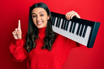 Young hispanic woman holding piano keyboard smiling with an idea or question pointing finger with...
