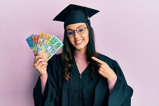 Young hispanic woman wearing graduation uniform holding australian dollars smiling happy pointing with hand and finger