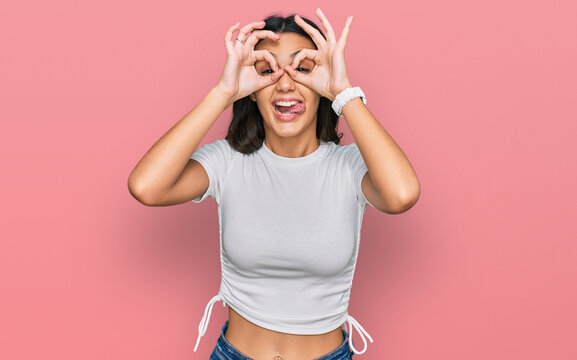 Young hispanic girl wearing casual white t shirt doing ok gesture like binoculars sticking tongue out, eyes looking through fingers. crazy expression.