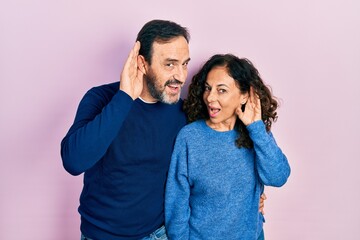 Middle age couple of hispanic woman and man hugging and standing together smiling with hand over ear listening an hearing to rumor or gossip. deafness concept. - obrazy, fototapety, plakaty