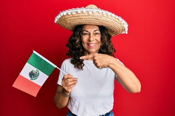 Middle age hispanic woman wearing mexican hat holding mexico flag smiling happy pointing with hand and finger