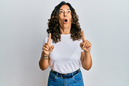 Middle age hispanic woman wearing casual white t shirt amazed and surprised looking up and pointing with fingers and raised arms.