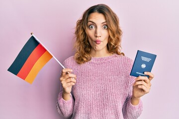 Young caucasian woman holding germany flag and passport making fish face with mouth and squinting...