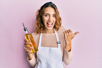 Young caucasian woman wearing apron holding olive oil can pointing thumb up to the side smiling...