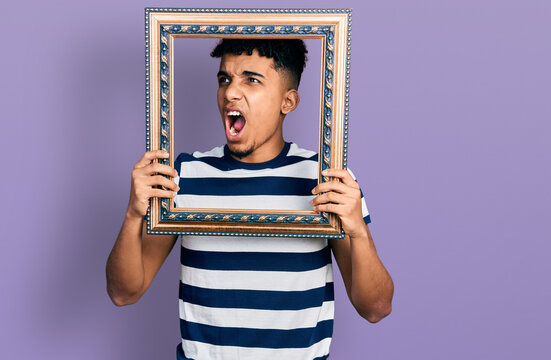 Young african american man holding empty frame angry and mad screaming frustrated and furious, shouting with anger. rage and aggressive concept.