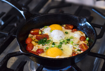 Breakfast eggs with tomatoes and green onions frying in cast iron pan on gas stove