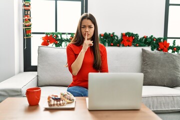Young brunette woman using laptop sitting on the sofa on christmas asking to be quiet with finger on lips. silence and secret concept.