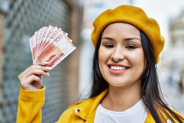 Young latin girl smiling happy holding norway krone banknotes at the city.