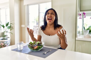 Obraz na płótnie Canvas Young hispanic woman eating healthy salad at home crazy and mad shouting and yelling with aggressive expression and arms raised. frustration concept.