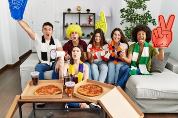 Group of young friends watching and supporting soccer match eating pizza at home.