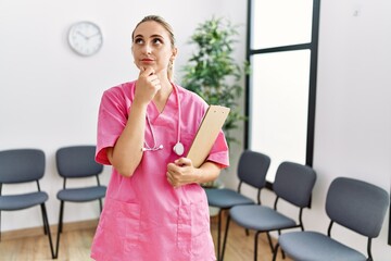 Young nurse woman at medical clinic waiting room serious face thinking about question with hand on...