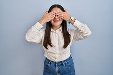 Young latin woman standing over blue background covering eyes with hands smiling cheerful and funny. blind concept.