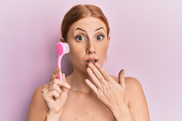Young irish woman using facial exfoliating brush covering mouth with hand, shocked and afraid for...