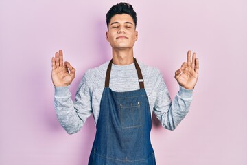 Young hispanic man wearing apron relax and smiling with eyes closed doing meditation gesture with fingers. yoga concept.