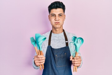 Young hispanic man wearing professional baker apron holding cooking tools puffing cheeks with funny face. mouth inflated with air, catching air.