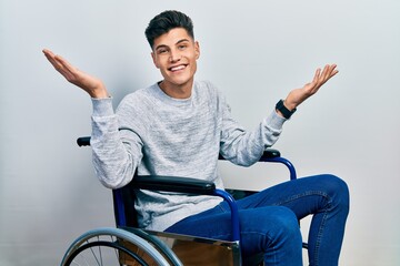 Young hispanic man sitting on wheelchair clueless and confused expression with arms and hands...
